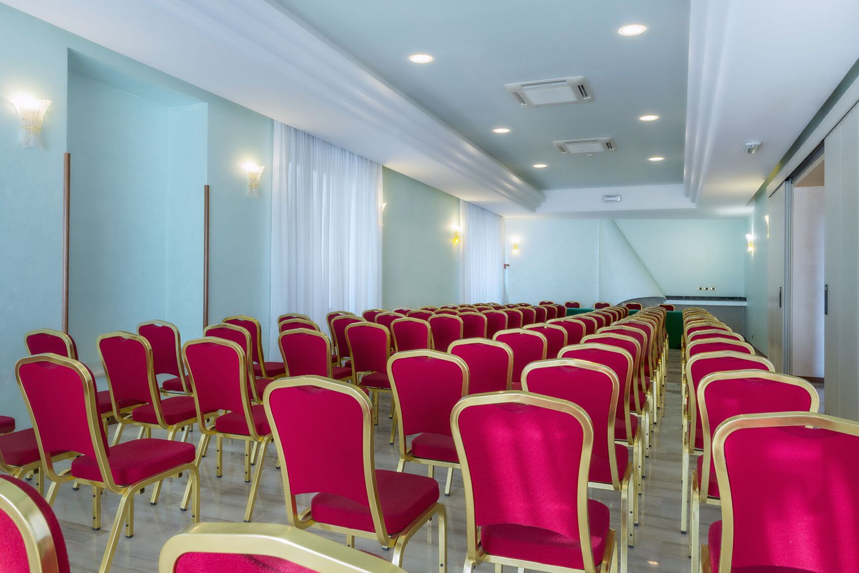 A conference room with rows of red chairs and gold frames, white curtains, and recessed ceiling lights.