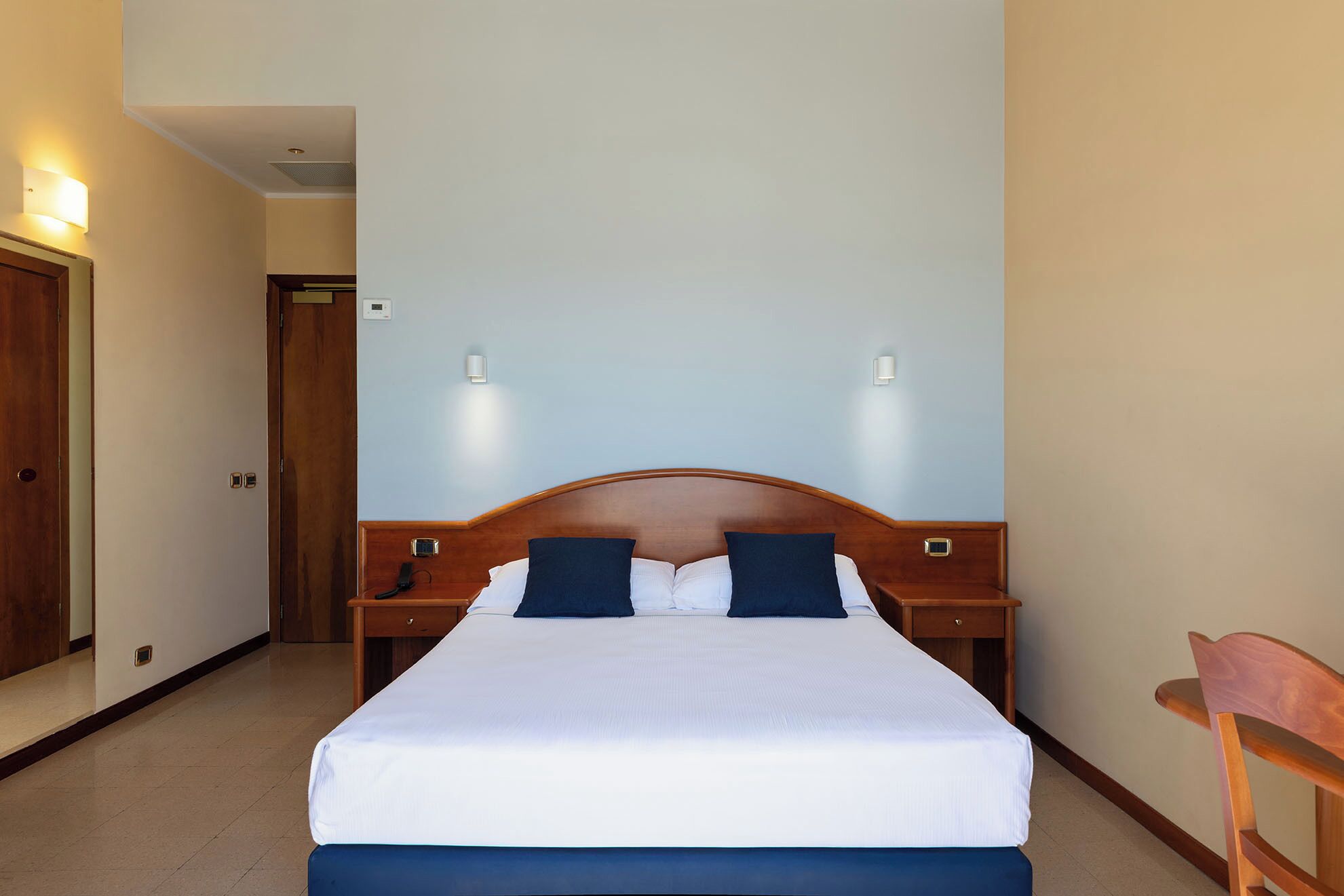 Une chambre d’hôtel avec un grand lit, deux tables de chevet, une chaise en bois et deux lampes fixées au mur.
