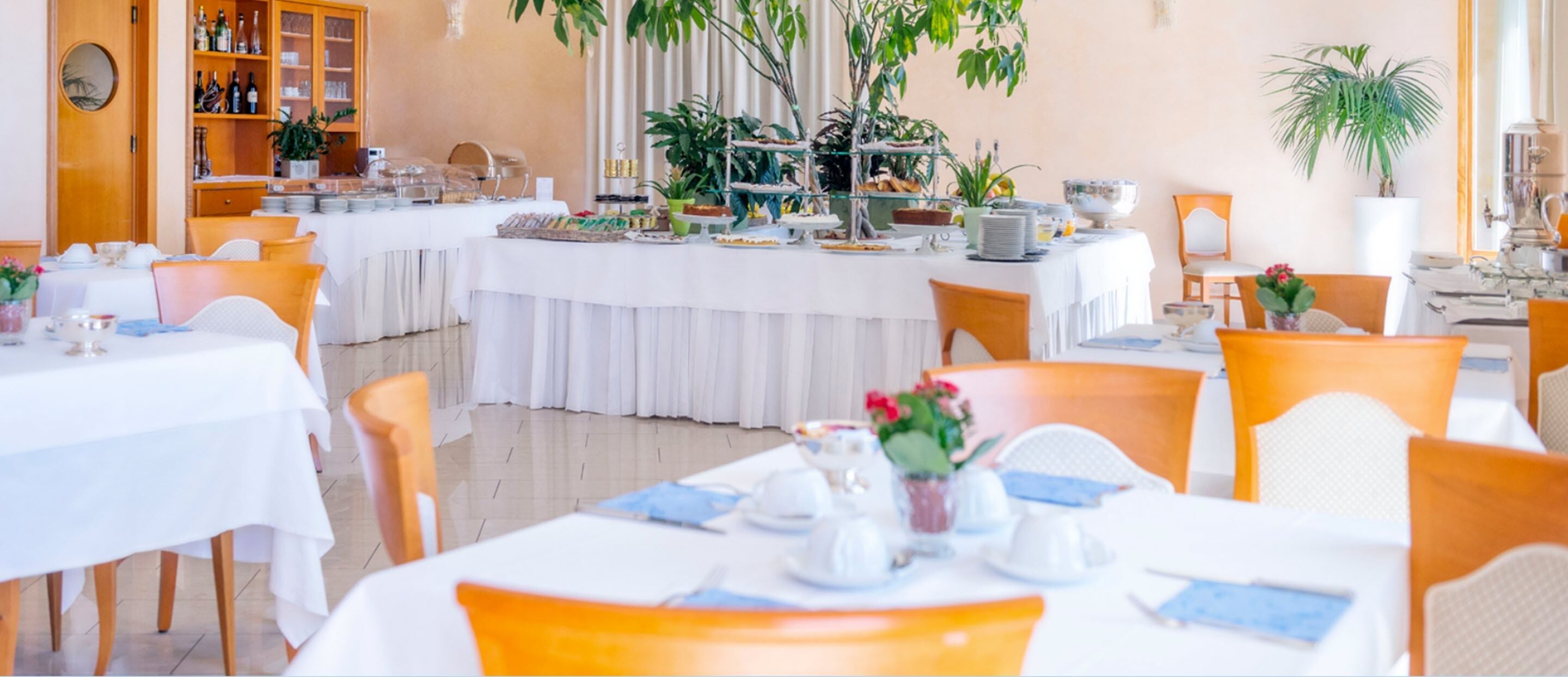 A restaurant with white tablecloths, orange chairs, and a buffet area with various dishes.