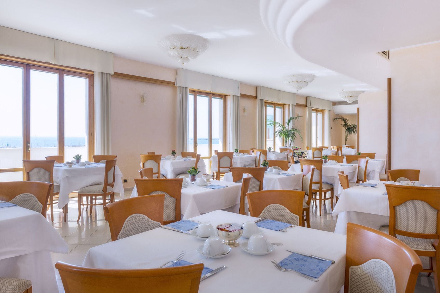 Une salle à manger avec des tables dressées pour le service, de grandes fenêtres et une vue sur la mer.