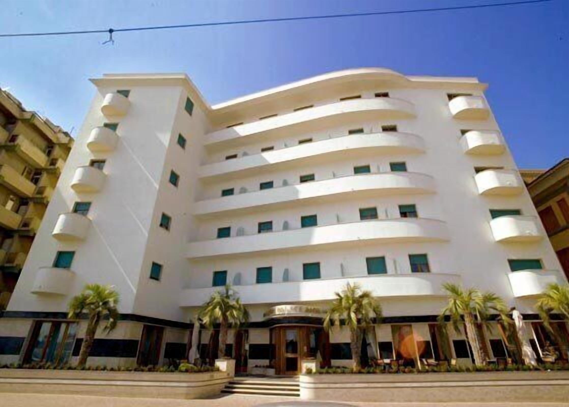 Un hotel a più piani con una facciata curva, balconi su ogni piano e palme di fronte.