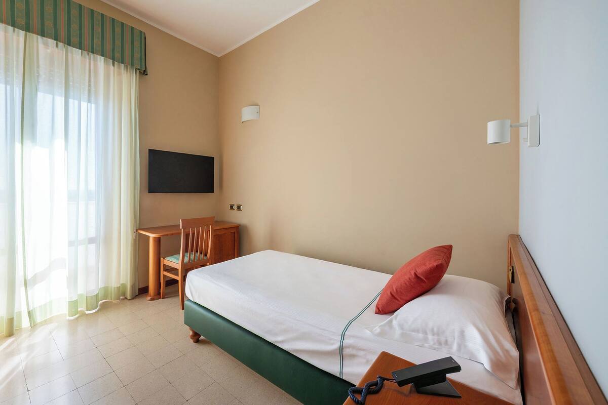 Quarto de hotel com cama, escrivaninha, cadeira, televisão e telefone.