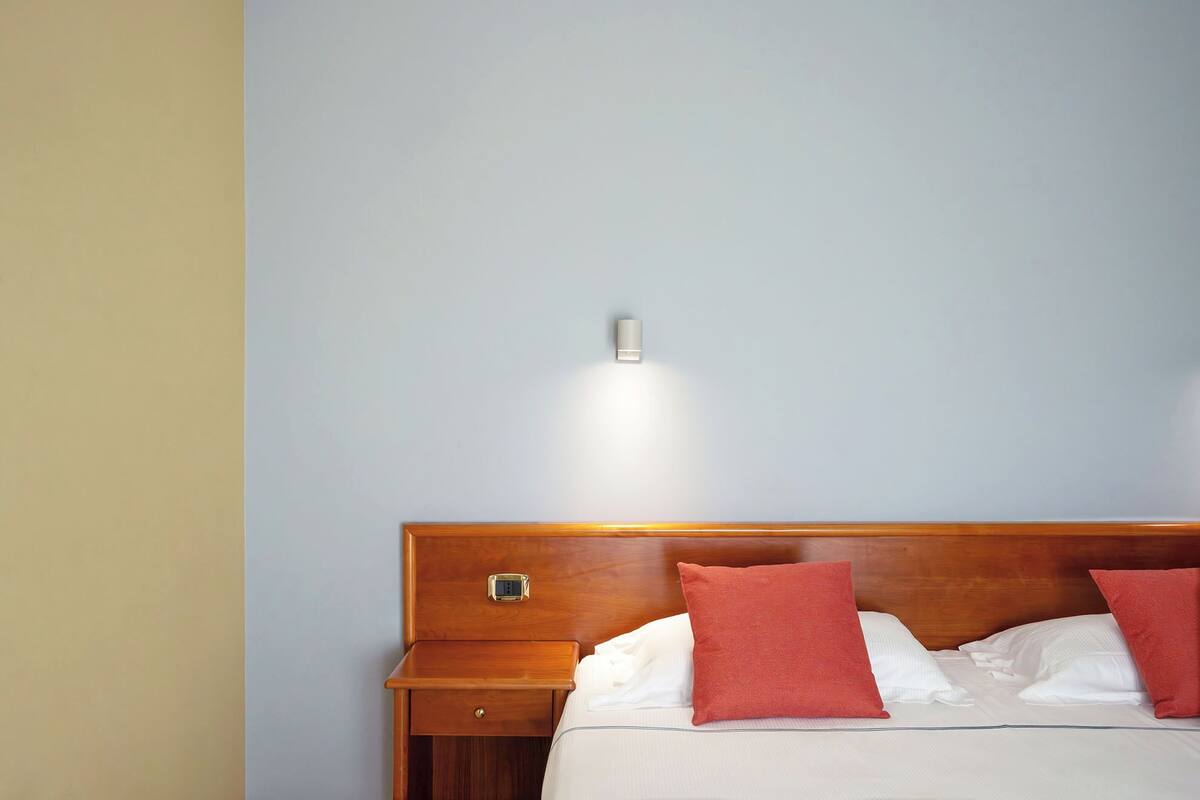 Quarto de hotel com cabeceira de madeira, mesa de cabeceira com gaveta e luminária fixa na parede.
