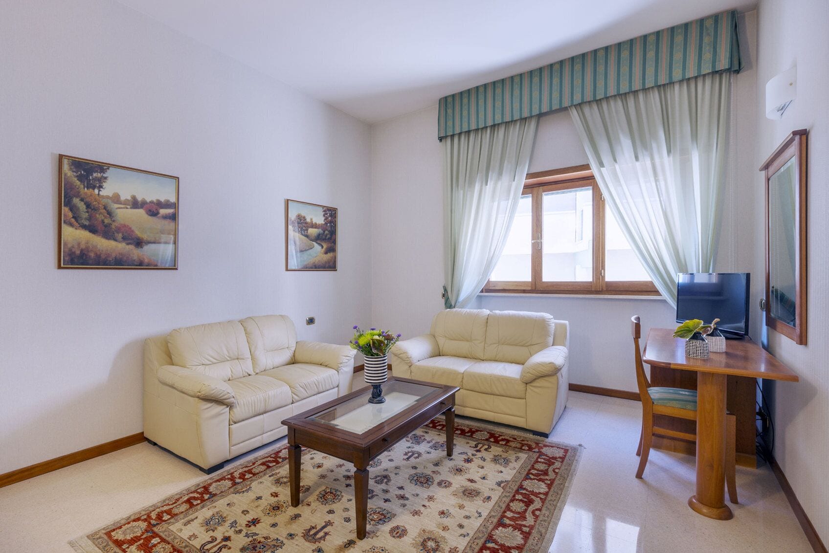 A living room with two cream-colored sofas, a wooden coffee table, a small wooden side table with a plant, a TV on a wooden stand, and a window with curtains.