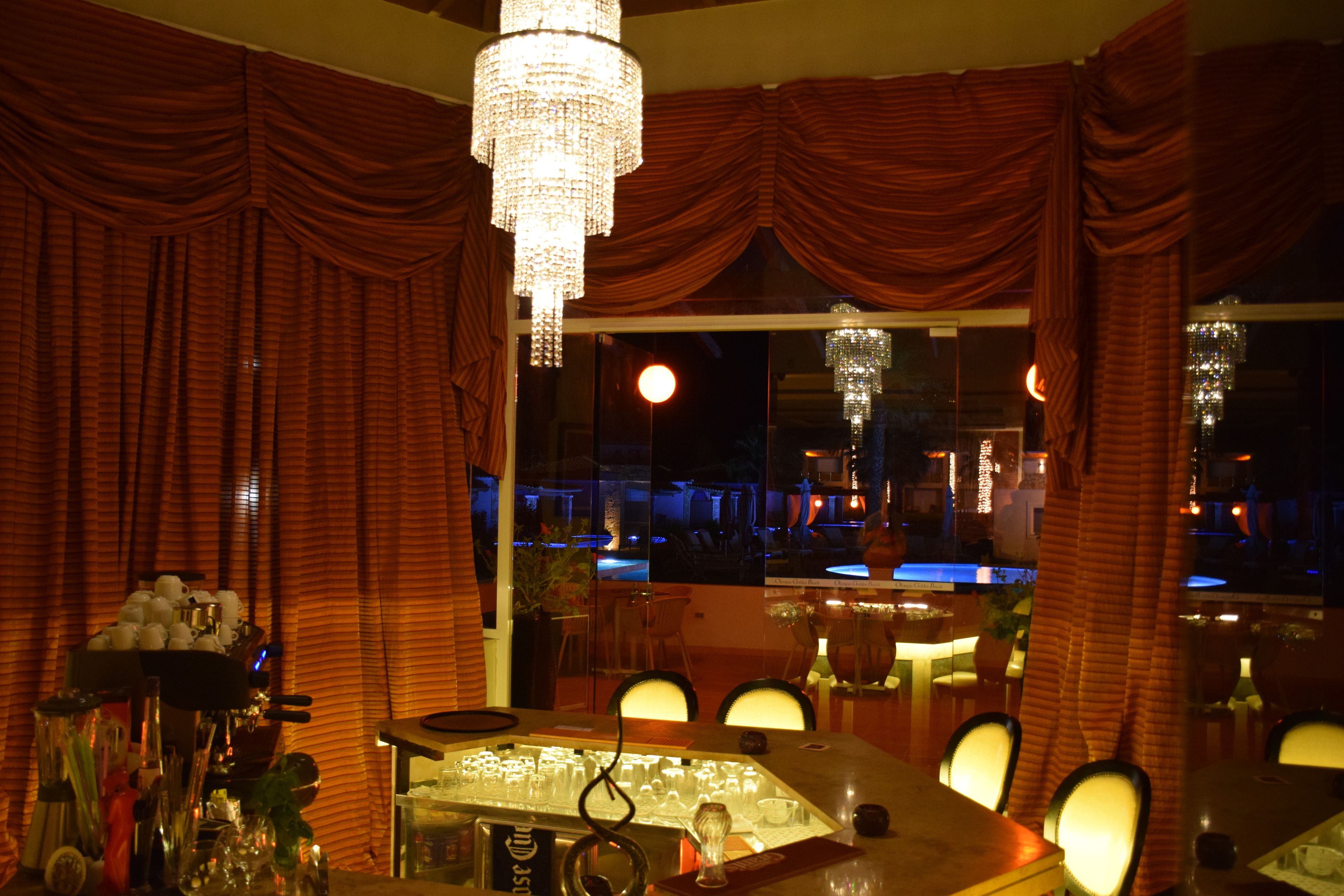 A dimly lit restaurant with red curtains, a bar area, and a view of the cityscape through large windows.