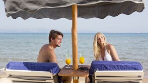 Two people sitting at a table with a beach view, under a canopy, with towels on chairs and drinks on the table. - Olympia Golden Beach & Spa (Andravida-Kyllini)