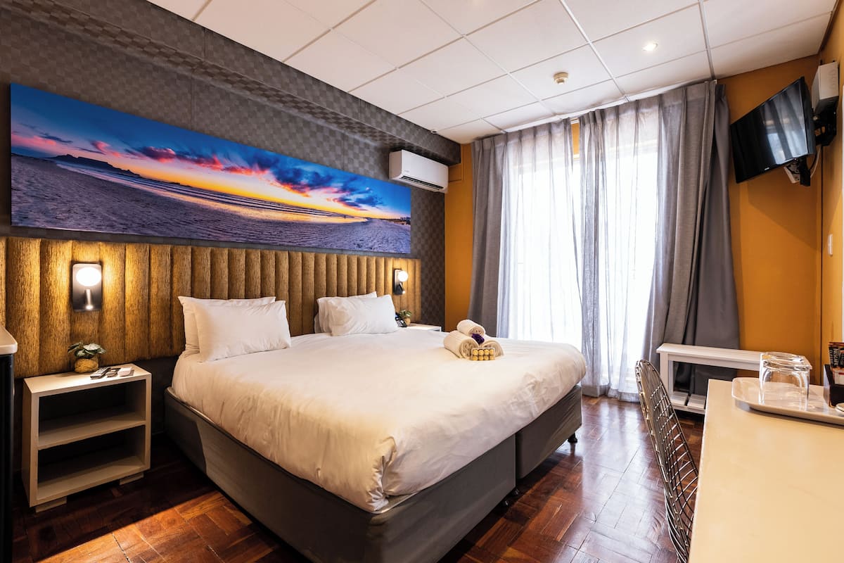 Ein Hotelzimmer mit einem großen Bett, einem flachbildfernseher an der Wand, einem Holzkopfteil und einem Gemälde, das einen Sonnenuntergang über dem Meer zeigt.