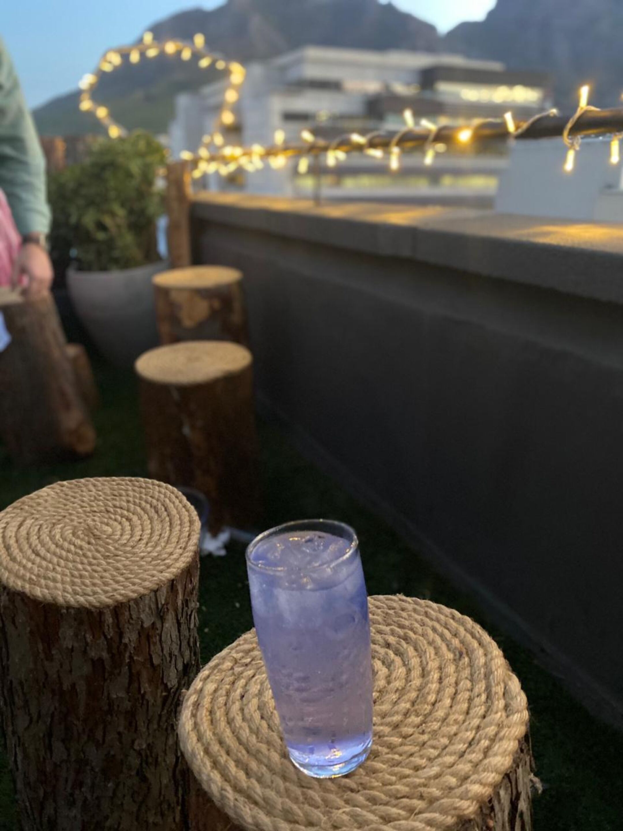 A glass of water on a woven stool with a view of a building and string lights in the background.