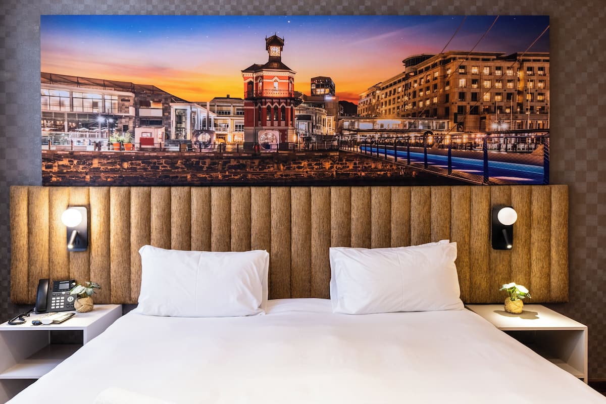 Ein Hotelzimmer mit großem Stadtbild über dem Bett, einem Holz-Kopfteil, zwei weißen Kissen, Nachttischen mit Lampen und einer kleinen Pflanze.