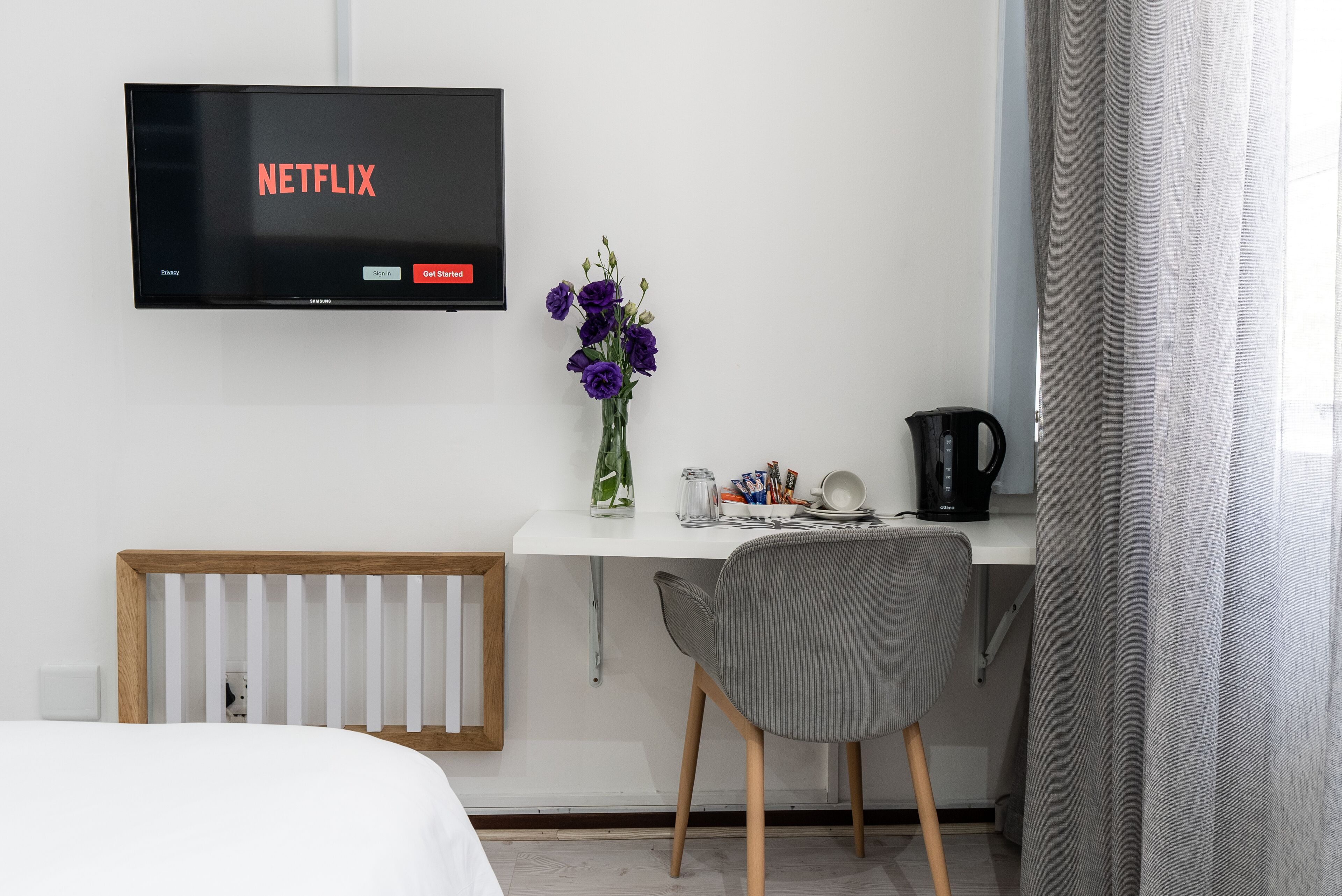 Habitación de hotel con cama, escritorio con silla, radiador, televisor mostrando 'NETFLIX', jarra con flores y cafetera.