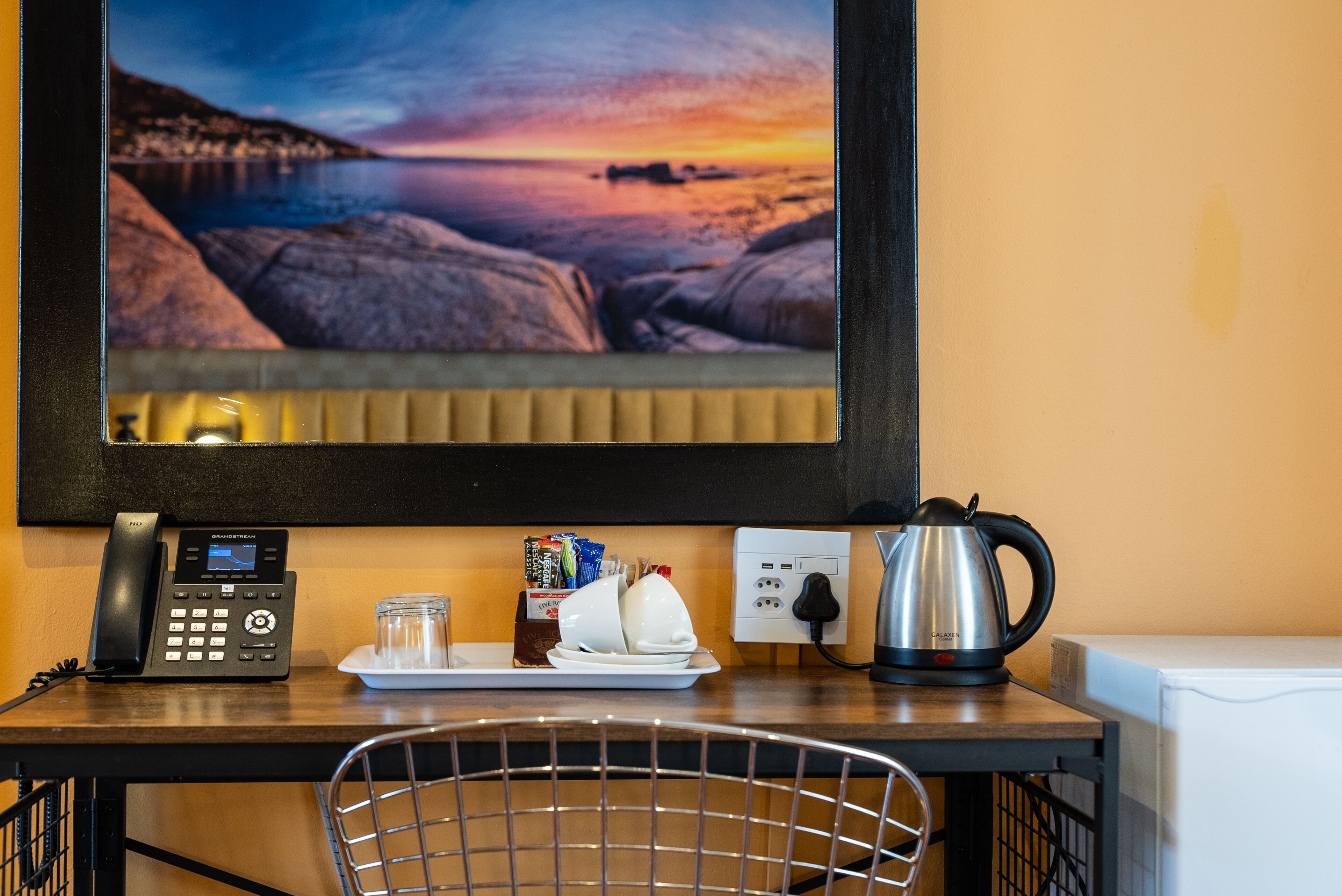 Et hotelværelse med udsigt til en solnedgang over havet, en telefon, en elkedel og et bakke med et glas vand og en vandflaske.