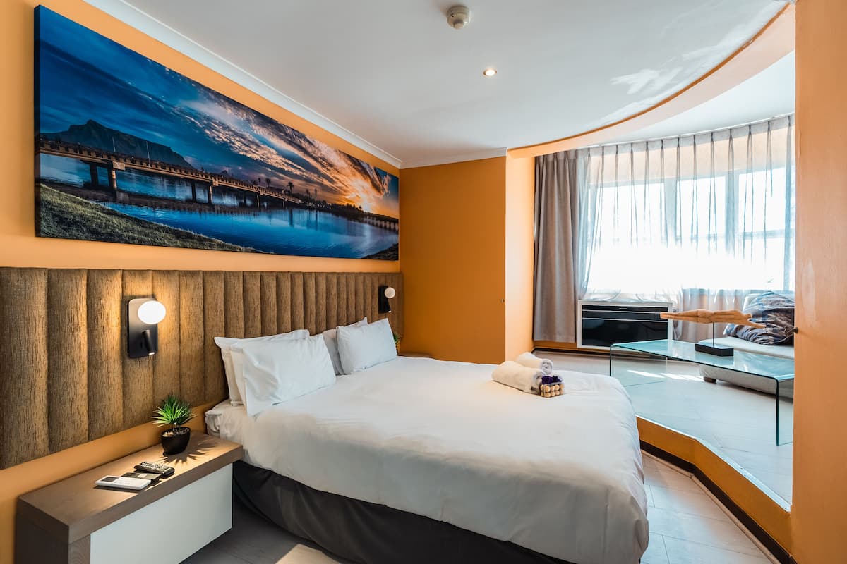 Ein Hotelzimmer mit einem großen Bett, einem Nachttisch mit Lampe, einem Gemälde einer Brücke und Blick ins Freie durch ein Fenster.