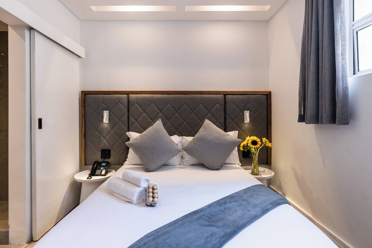 Ein modernes Hotelzimmer mit einem großen Bett, einem Nachttisch mit Telefon, einer Vase mit Sonnenblumen und einer blauen Tagesdecke.