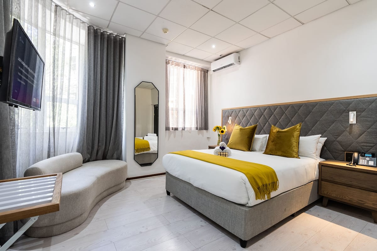 Ein modernes Hotelzimmer mit einer gebogenen Couch, einem großen Bett mit gestepptem Kopfteil, einem Nachttisch mit Lampe und einem an der Wand montierten Flachbildfernseher.
