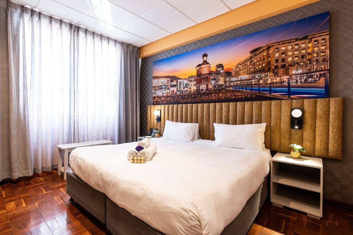 Ein Hotelzimmer mit einem großen Bett, einem Nachttisch, einer Wandlampe und einem Stadtbild an der Wand.