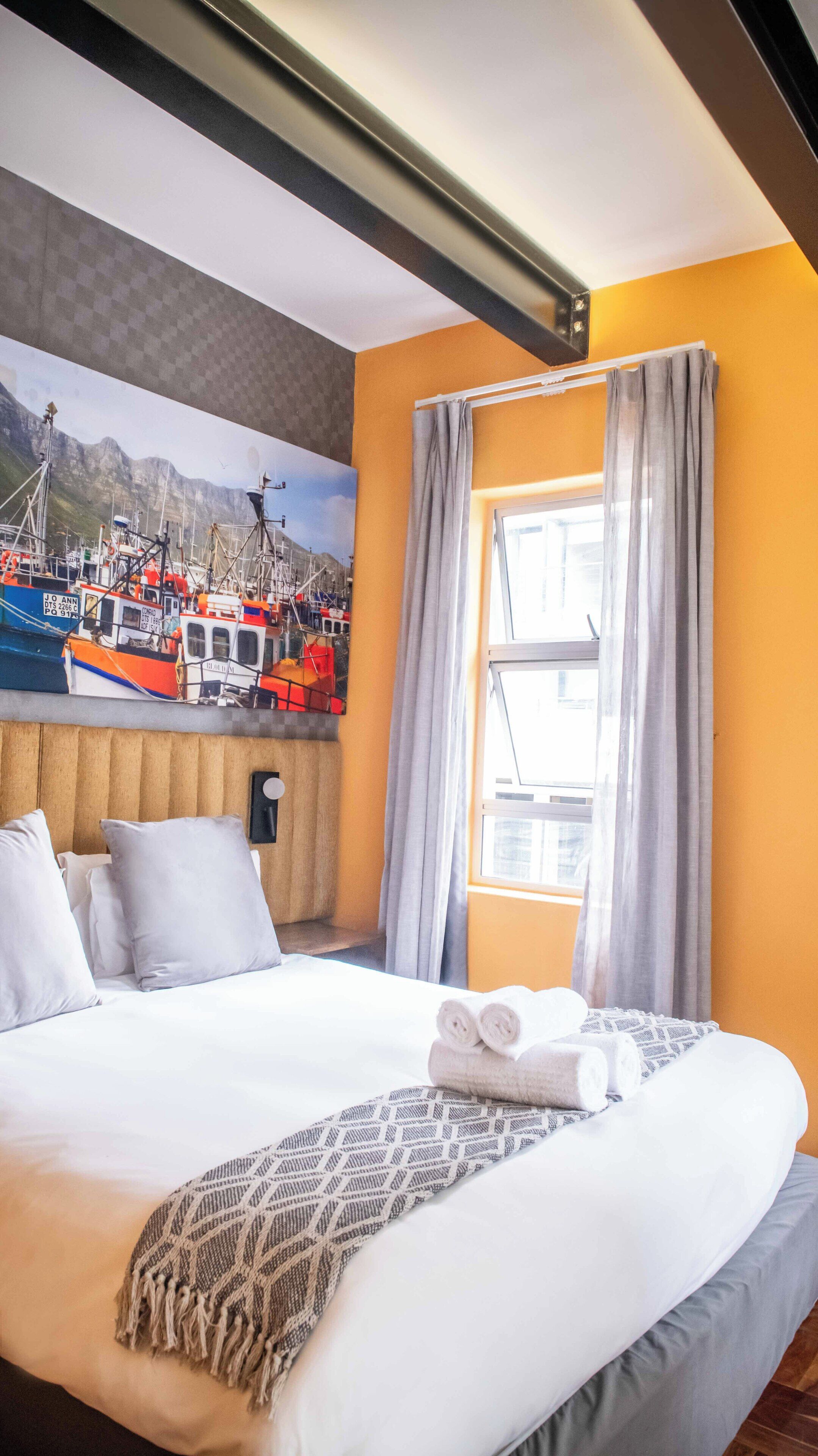 A hotel room with a bed, pillows, a blanket, a window with curtains, and a mural of boats on the wall.