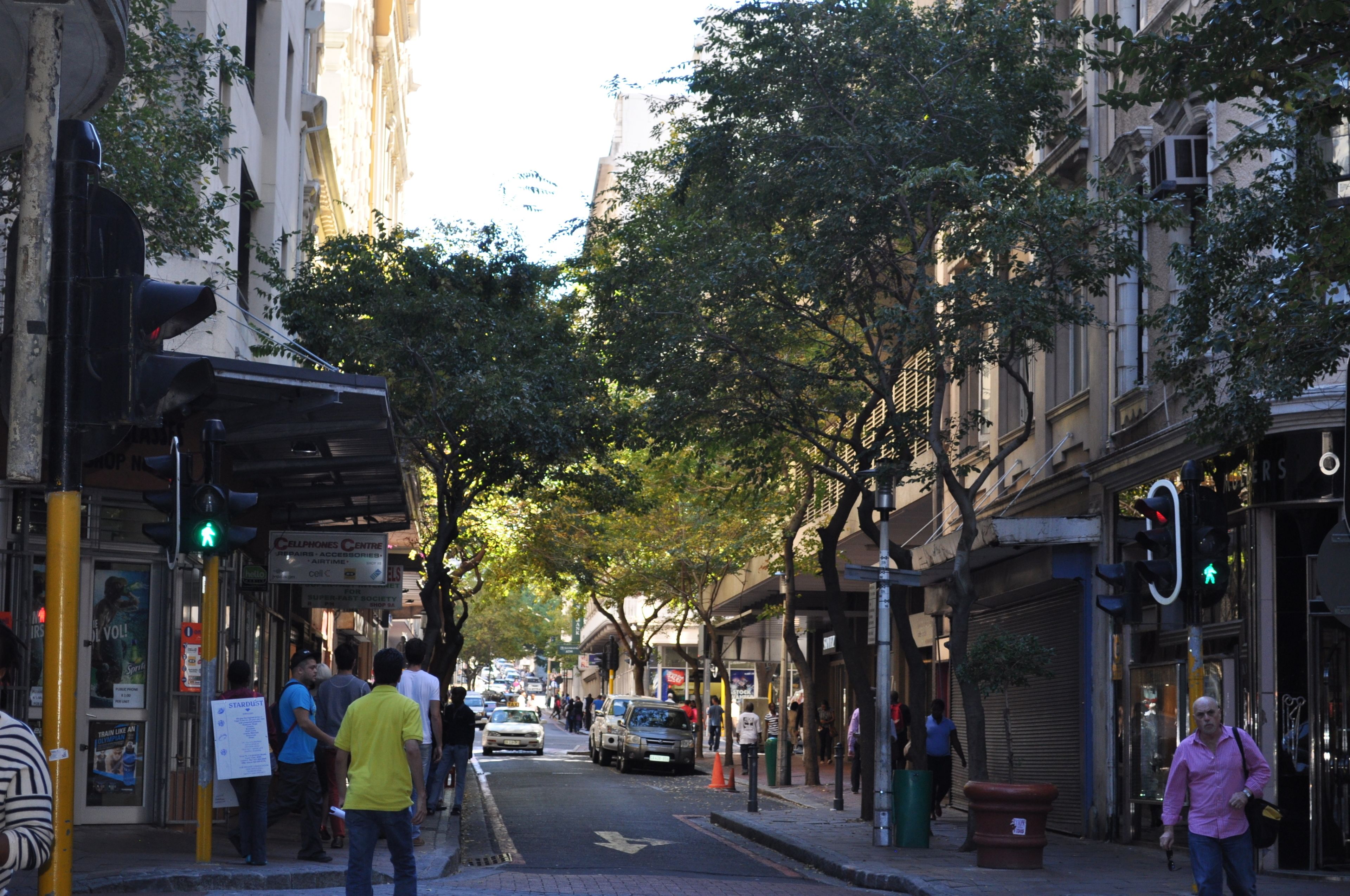 A street lined with shops and trees, with pedestrians and vehicles.