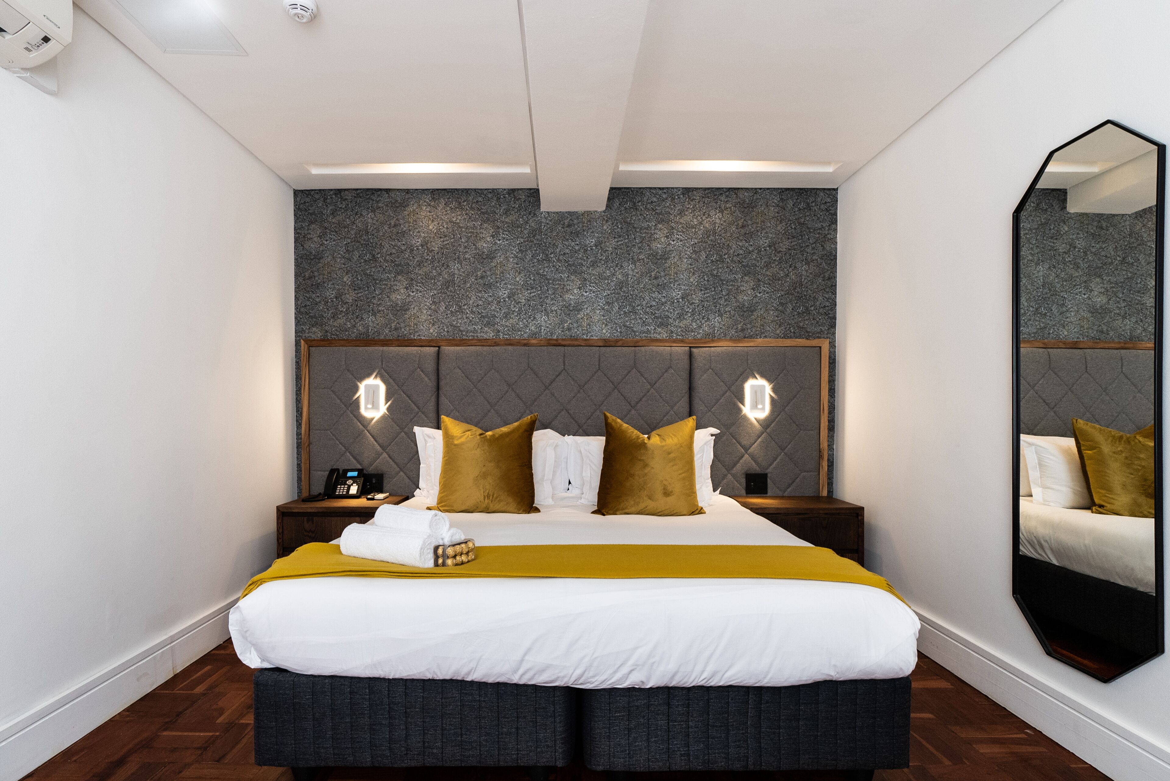 Une chambre d’hôtel moderne dotée d’un grand lit, d’une tête de lit avec éclairage intégré, d’un miroir et d’un plancher en bois.