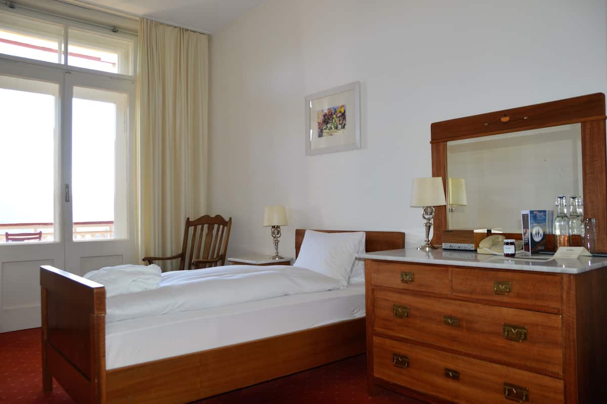 Ein Hotelzimmer mit einem hölzernen Bett, einer Kommode mit Schubladen, einem Spiegel, einem Sessel, einem Tisch mit Lampe und einem Fenster mit Vorhängen.