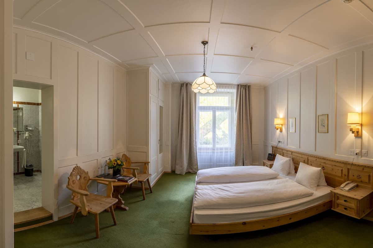Ein Hotelzimmer mit einem großen Bett, einem Schreibtisch mit Stuhl, einem kleinen Tisch mit Blumenvase sowie einem Nachttisch mit Lampe und einer Tür.