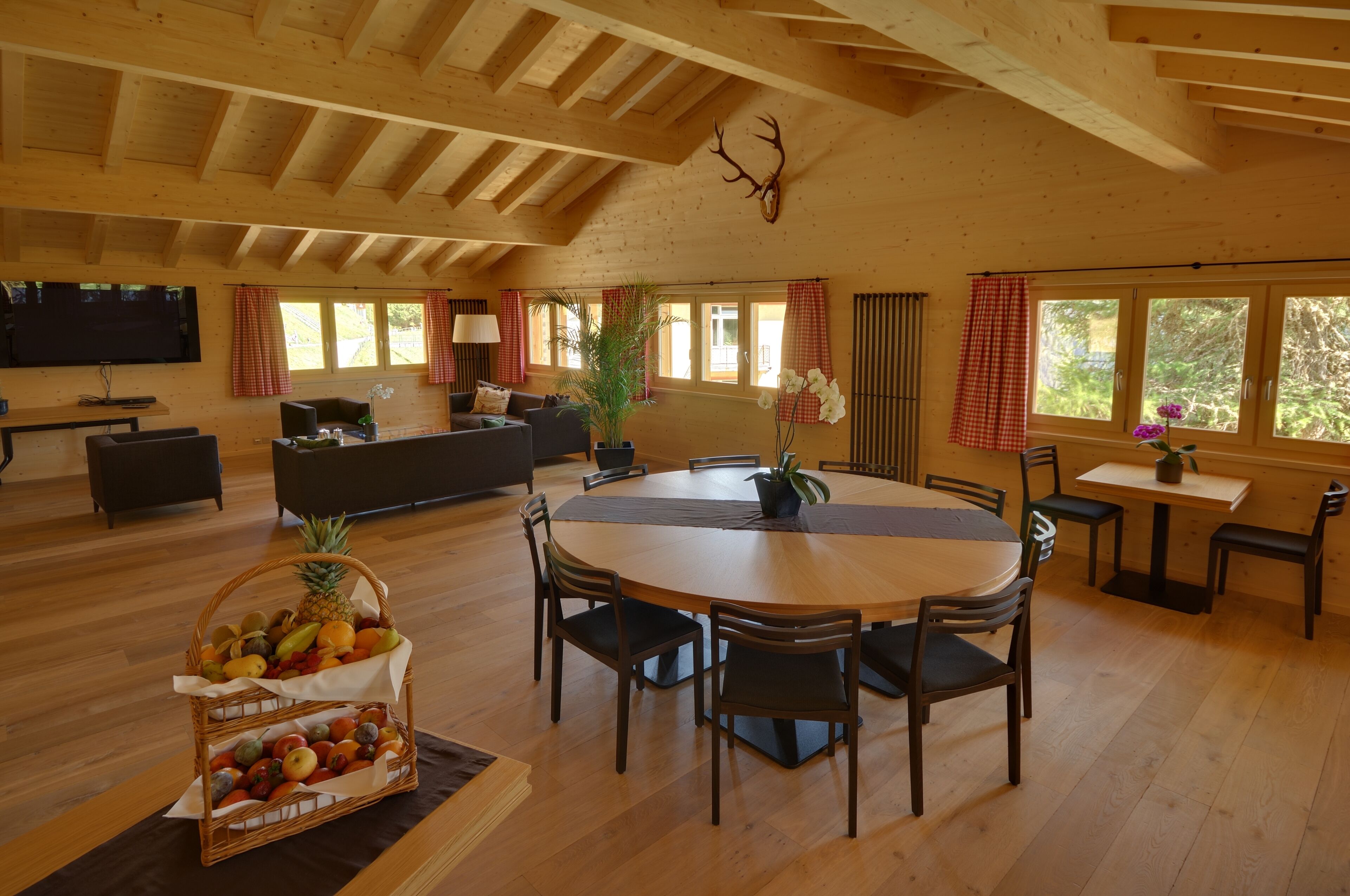 A spacious living room with a wooden ceiling, a round dining table surrounded by chairs, a television mounted on the wall, and a large window with curtains.