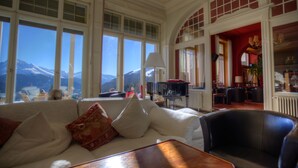 A spacious living room with a white sofa, a wooden coffee table, and a black armchair. Large windows offer a view of snow-capped mountains. - Schatzalp Snow & Mountain Resort (Davos)