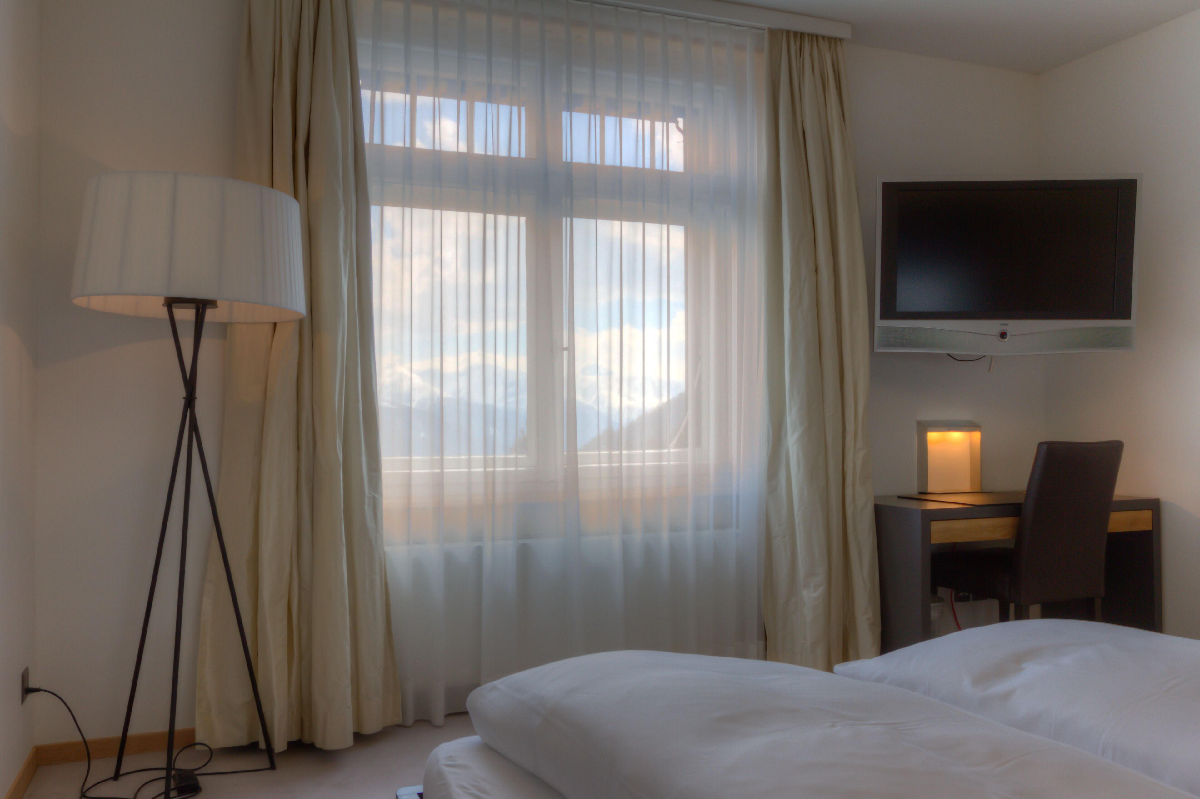 Une chambre d’hôtel comprenant un lit, un bureau avec une chaise, un téléviseur fixé au mur et une lampe sur pied.