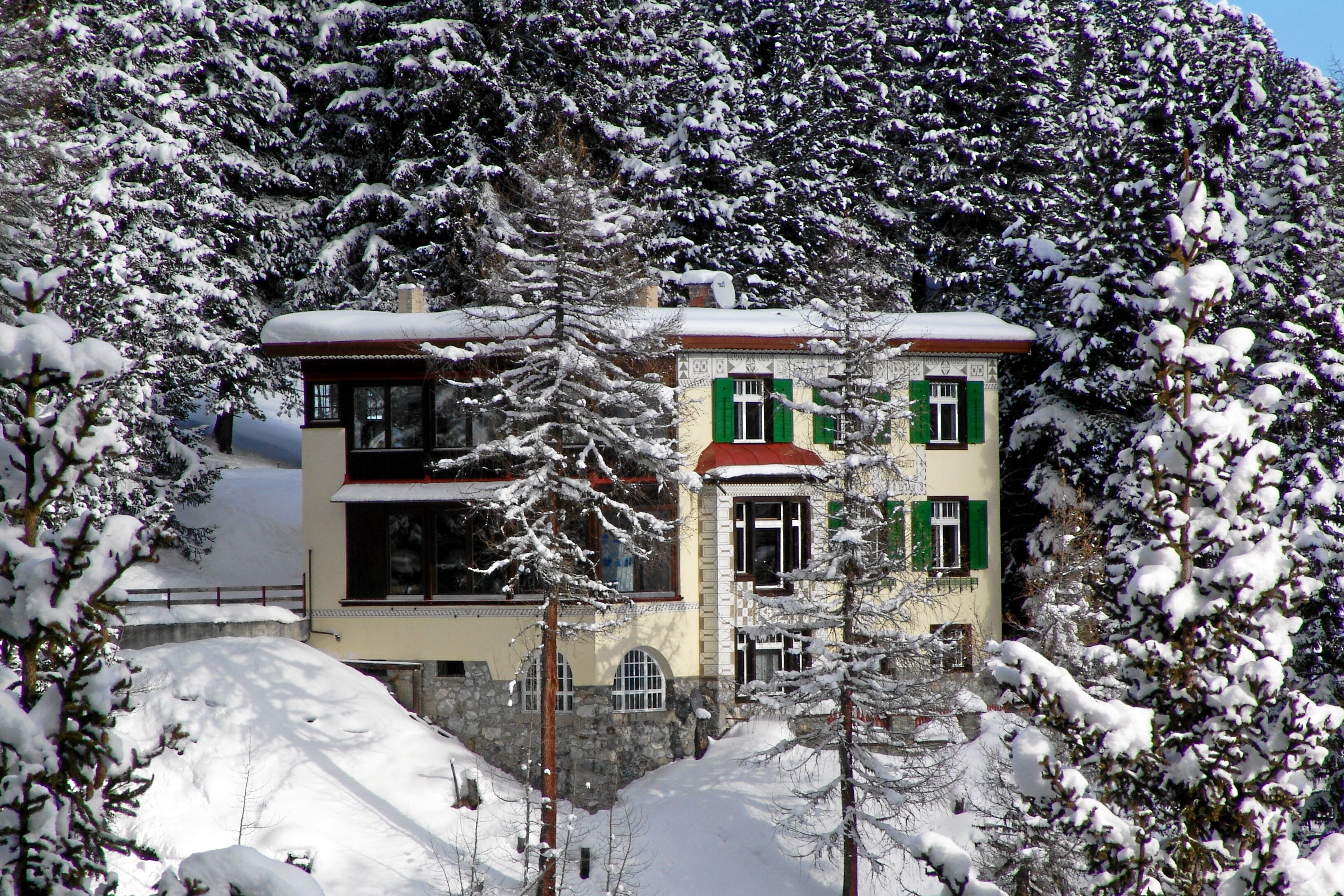 A two-story building with a snow-covered roof and green shutters, surrounded by snow-laden trees.