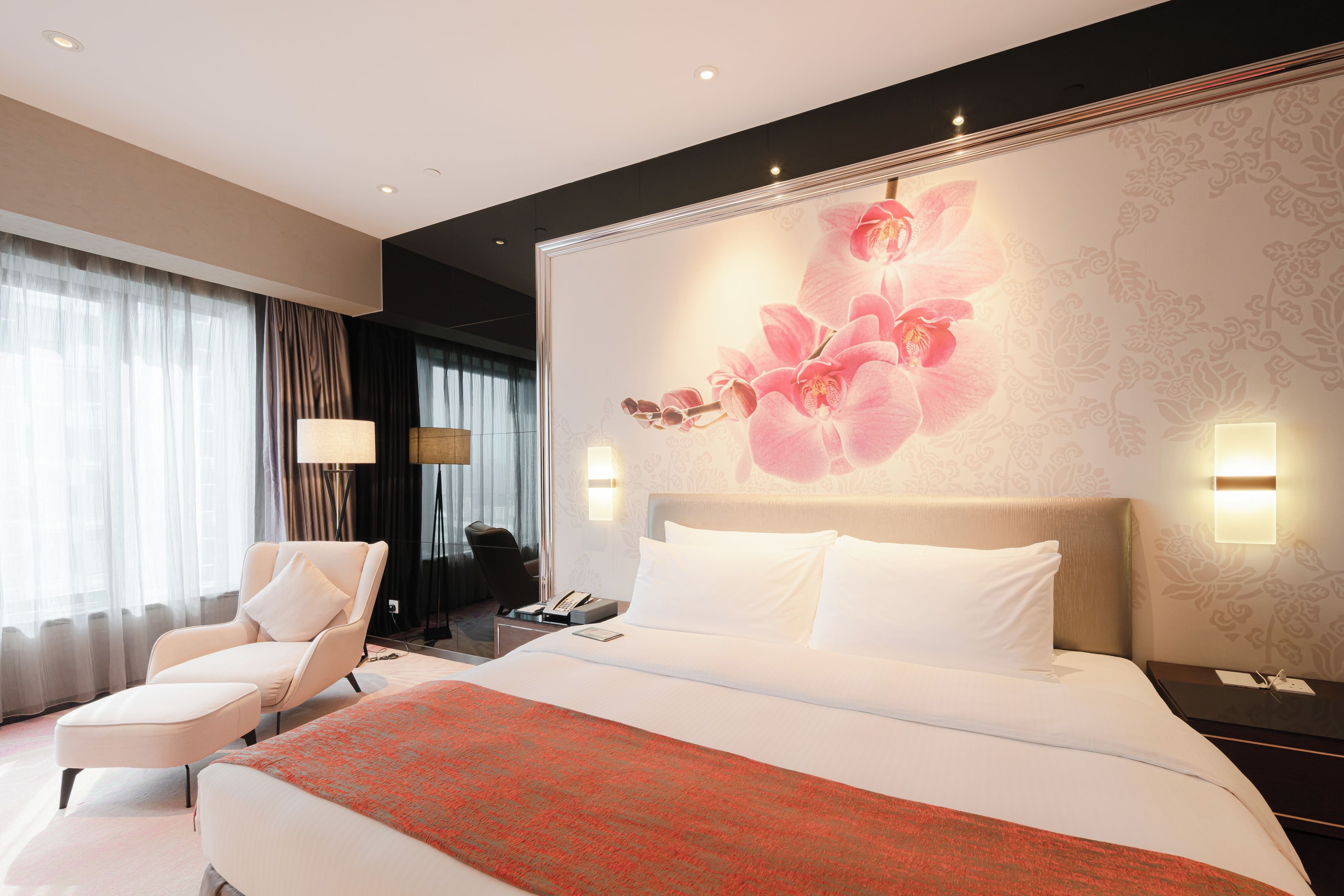 A hotel room with a large bed, a chair, a desk with a phone, and a wall with a floral design.