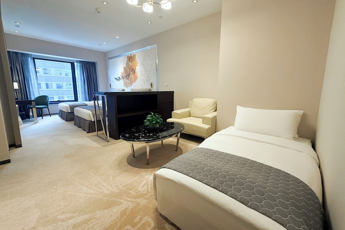 A modern hotel room with a large bed, a seating area with a sofa and armchairs, a small coffee table, and a television.