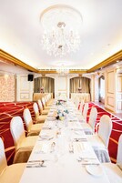 A banquet hall with a long table set for a formal event, surrounded by chairs with white covers, a chandelier, and a red carpet.