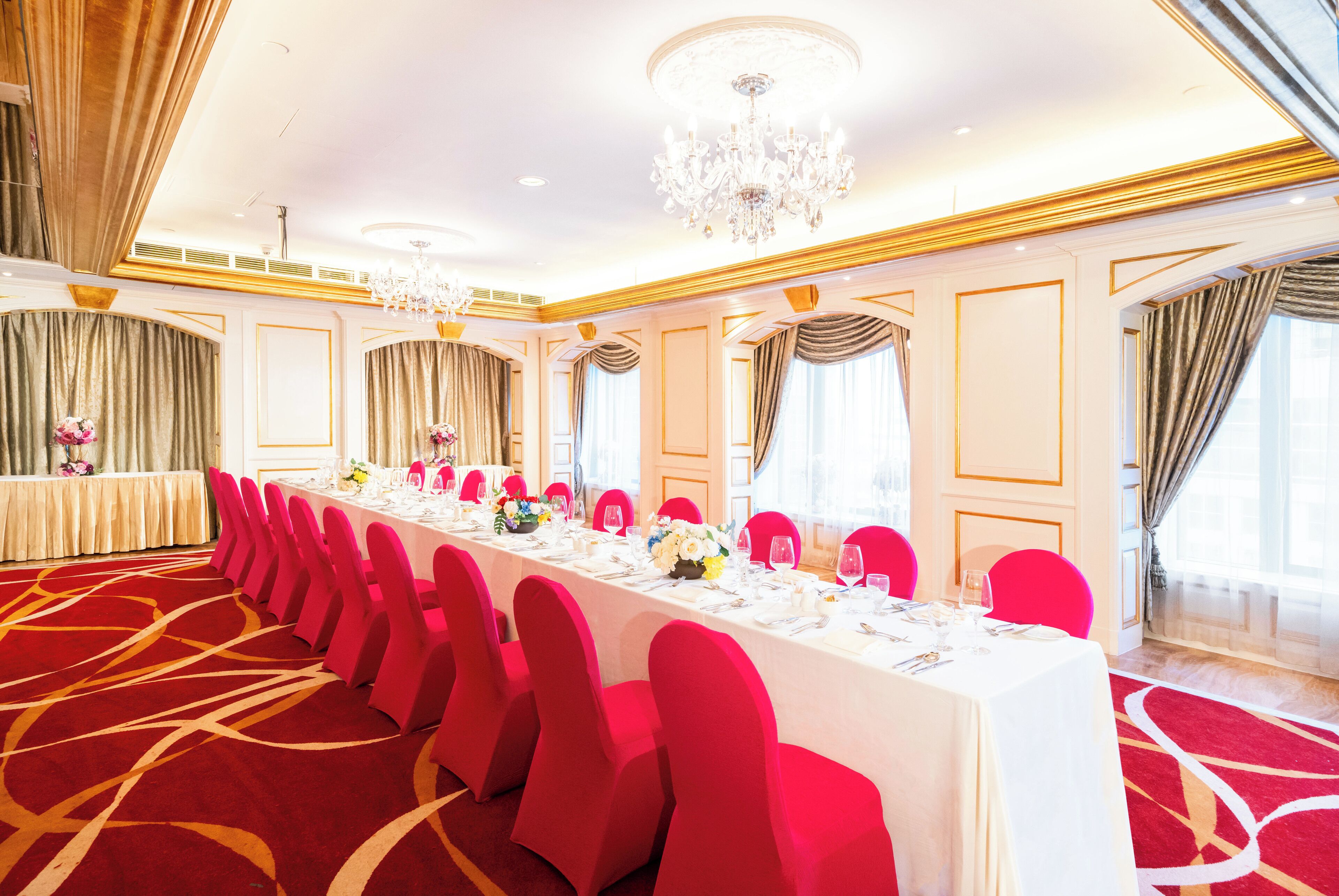 A banquet hall with a long table, red chairs, and chandeliers.