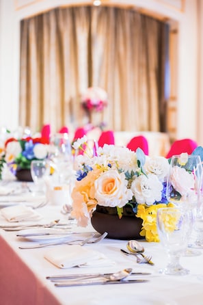 A long dining table set with white tablecloths, silverware, and glassware, with floral centerpieces in the foreground.
