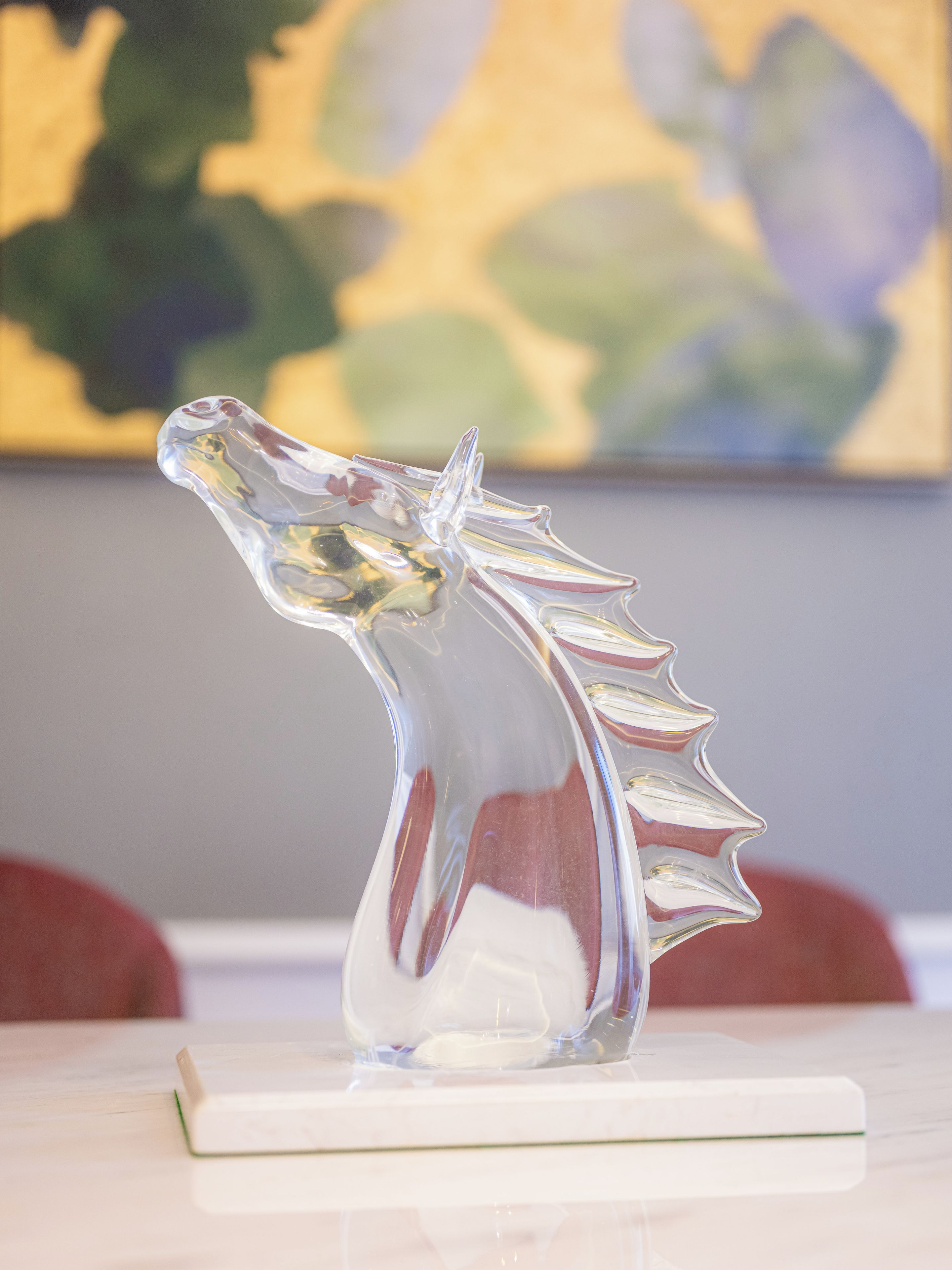 A clear glass sculpture of a horse's head on a white base, placed on a table with a blurred painting in the background.