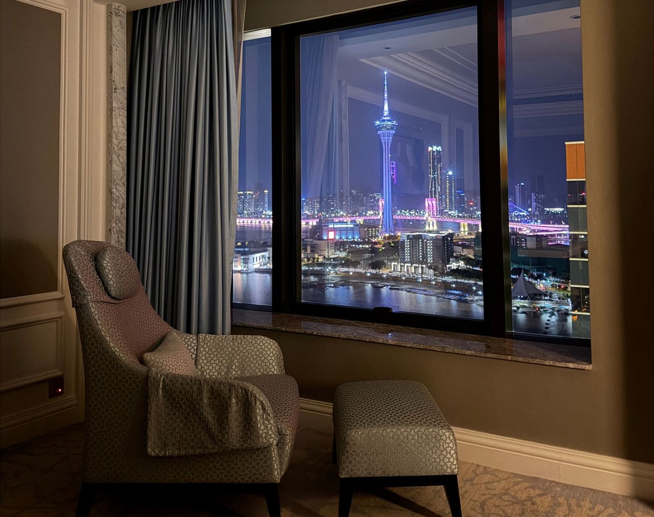 Una habitación con un ventanal grande que da a un paisaje urbano nocturno, con una silla y un reposapiés.