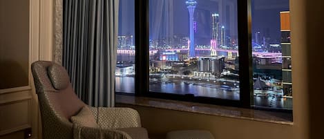 Una habitación con un ventanal grande que da a un paisaje urbano nocturno, con una silla y un reposapiés.