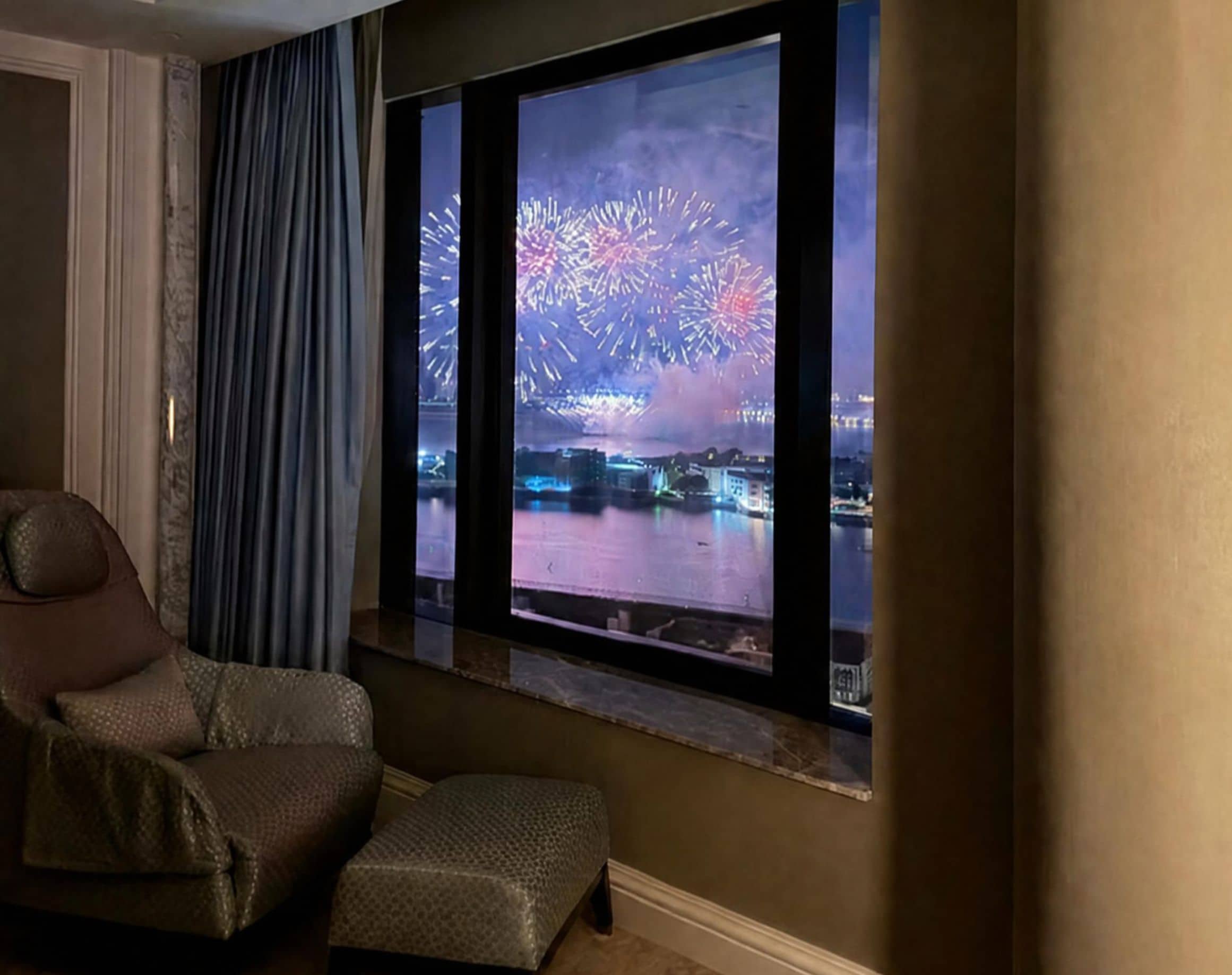 A room with a large window displaying fireworks over a body of water.