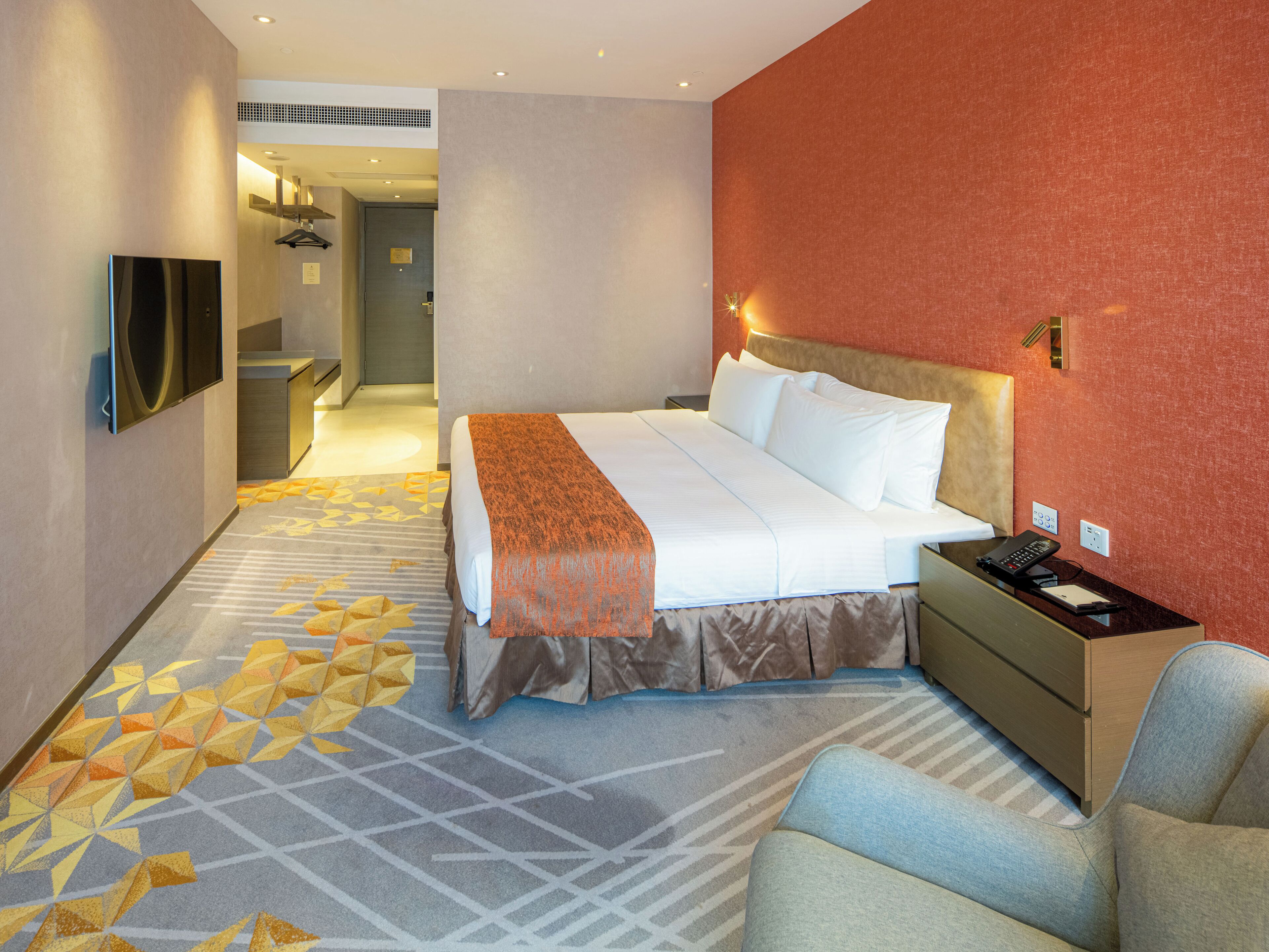 Habitación de hotel con cama, mesita de noche, silla, televisor y alfombra estampada.