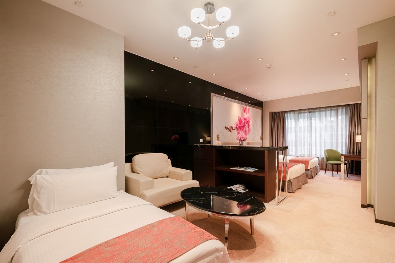 A hotel room with a bed, a sofa, a desk, and a chair. There is a painting on the wall and a window with curtains.