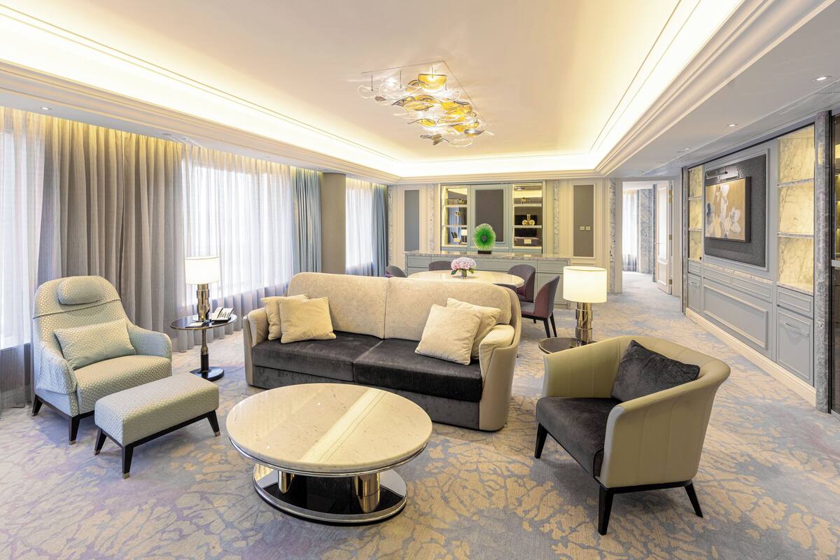 A spacious hotel room with a large sofa, armchairs, a round coffee table, and a dining area with a chandelier.