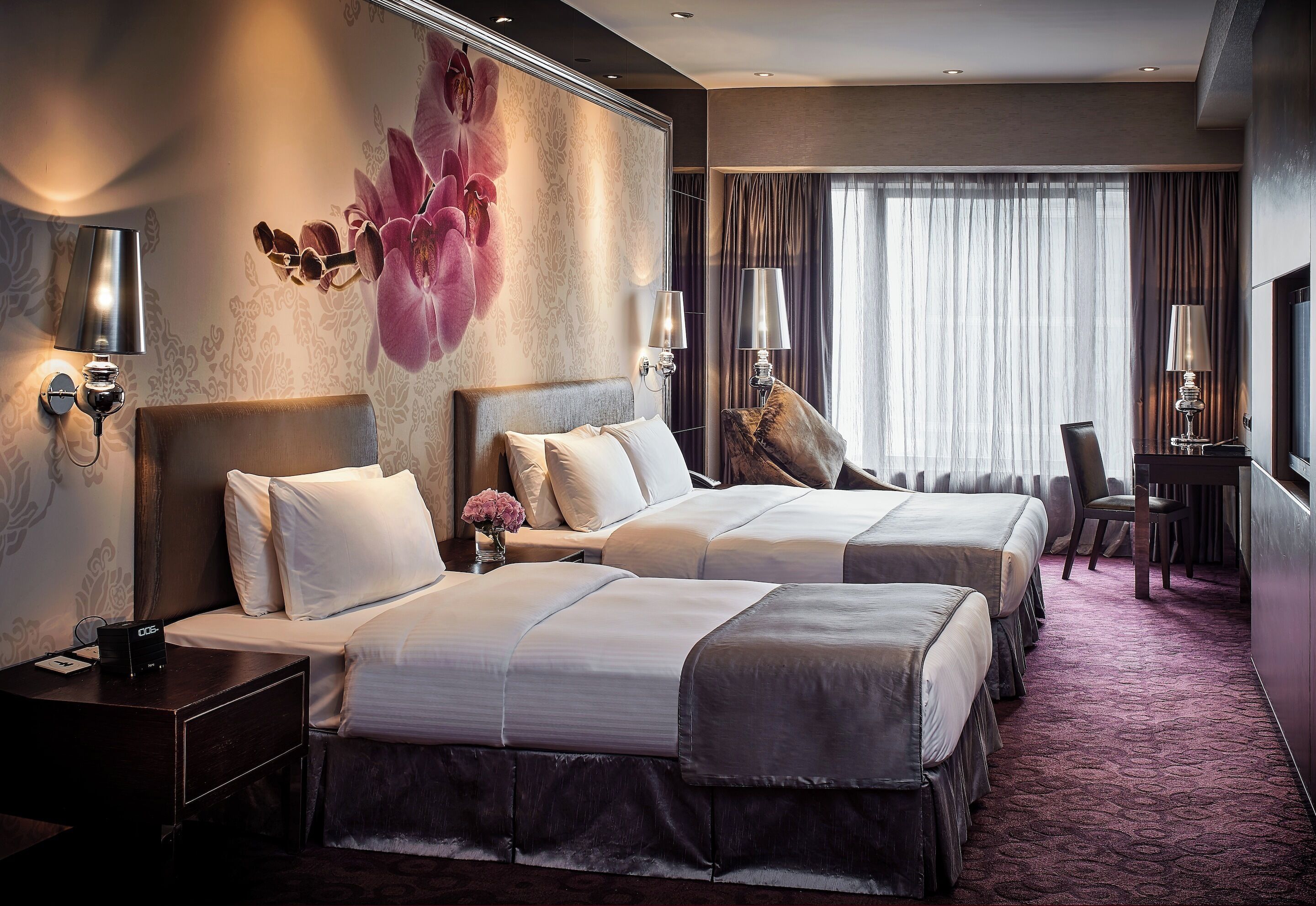 Ein Hotelzimmer mit zwei Betten, einem Schreibtisch und einem Sessel. Es gibt Wandleuchten und ein großes Fenster mit Vorhängen.