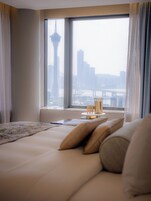 Habitación de hotel con un gran ventanal que ofrece vista panorámica de la ciudad, una cama con almohadas y una mesita de noche con una lámpara y un libro.