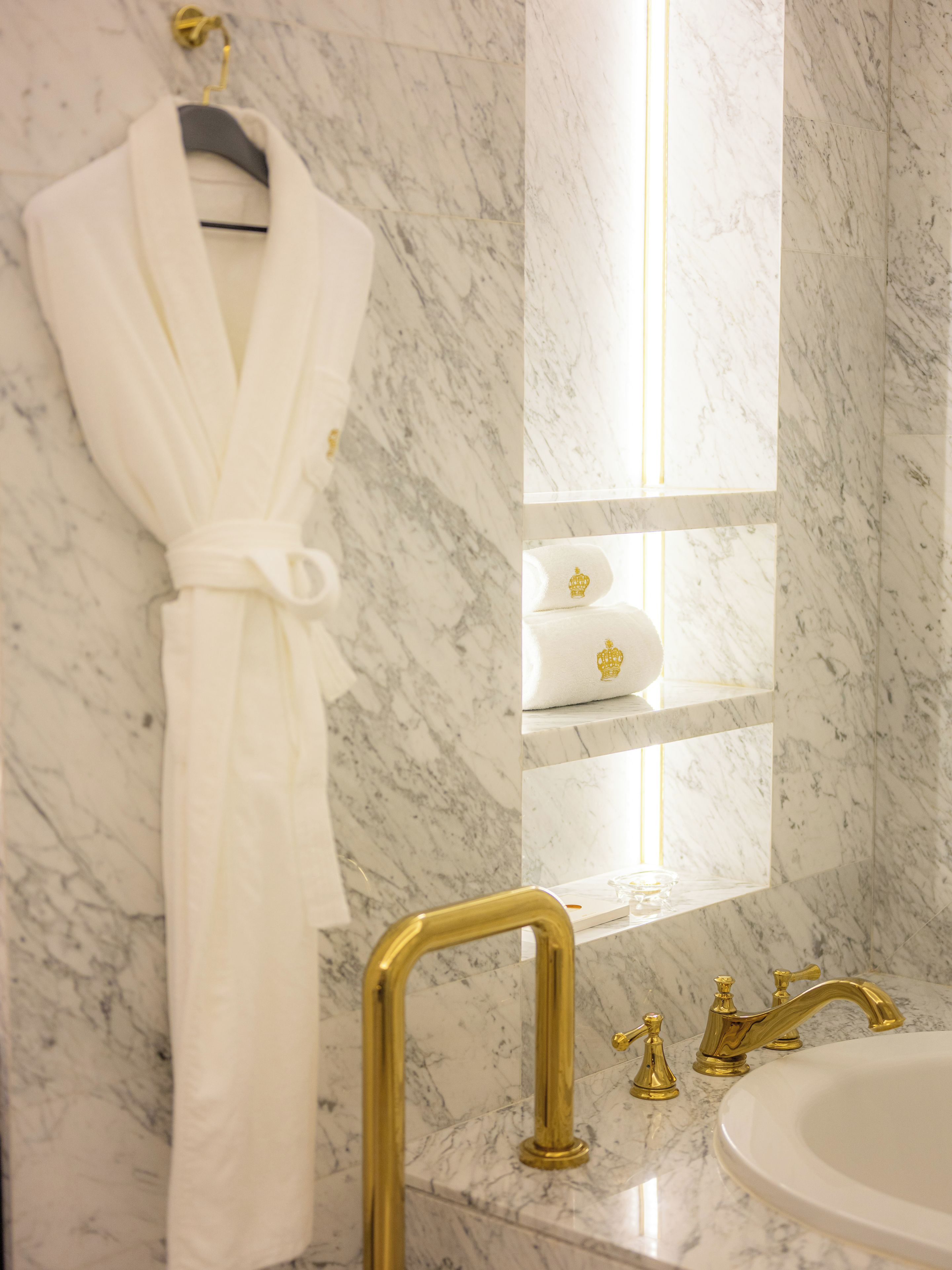 A bathroom with a marble floor, a white robe hanging on a rack, a gold faucet, and a mirror with shelves.