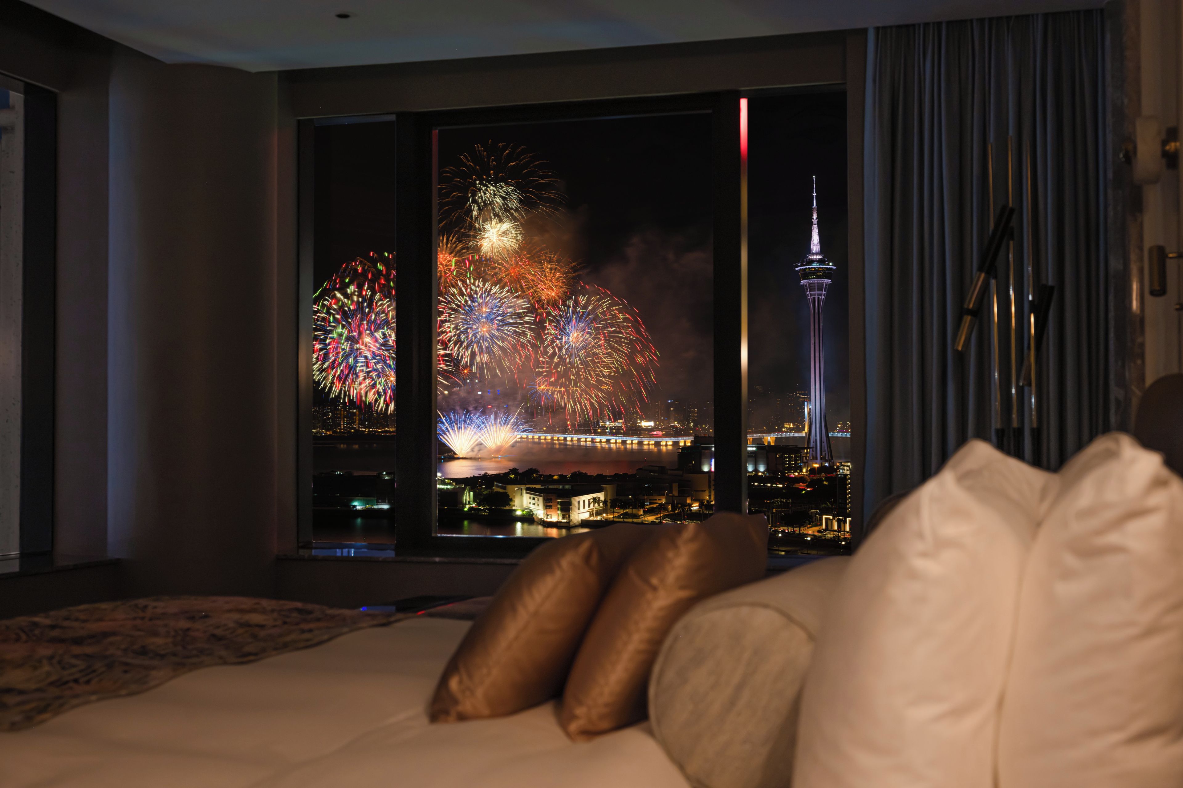 Ein Hotelzimmer mit großem Fenster und Blick auf Stadtlandschaft sowie Feuerwerk.