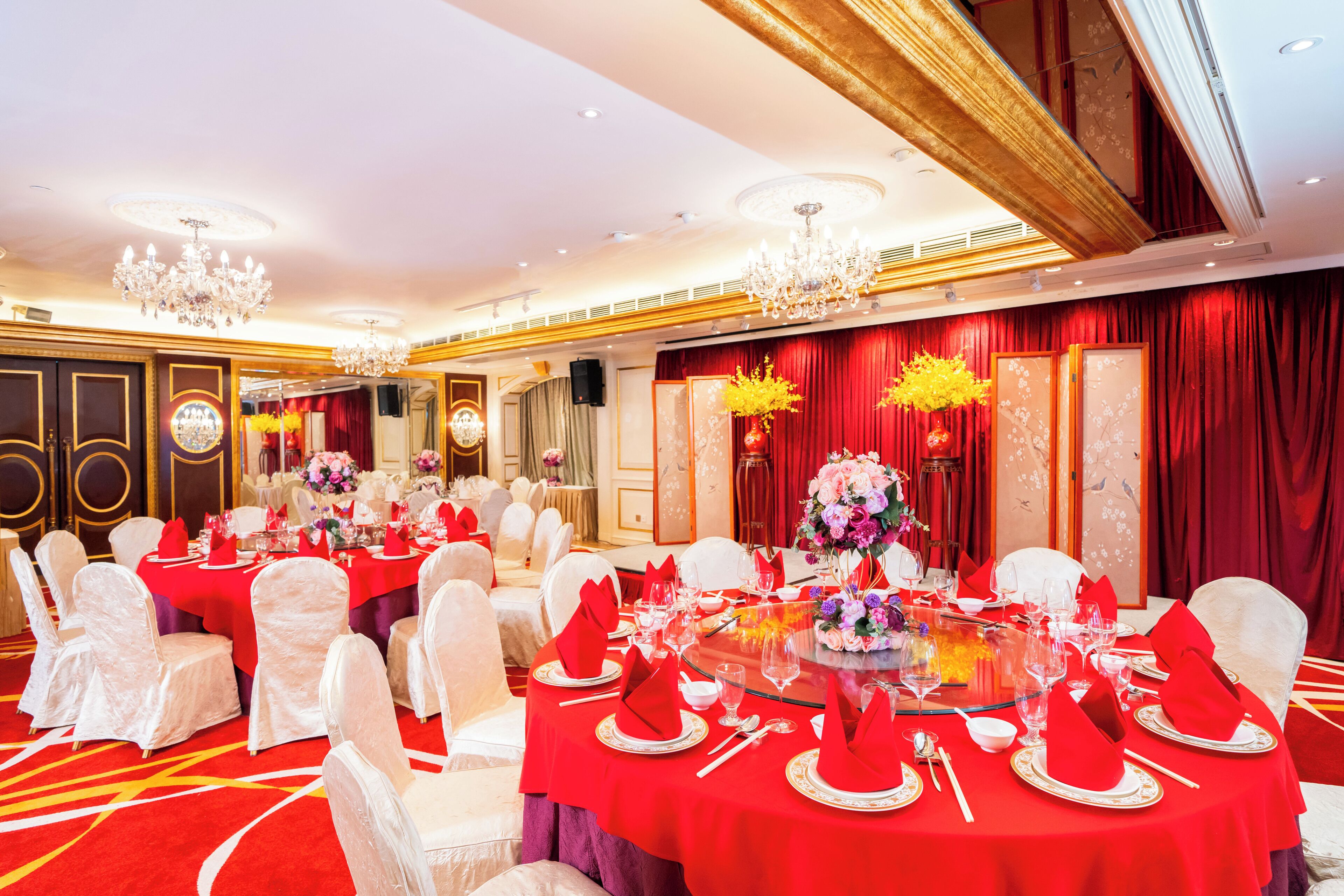Un salón de banquetes con mesas redondas dispuestas para un evento formal, con manteles rojos y sillas blancas.