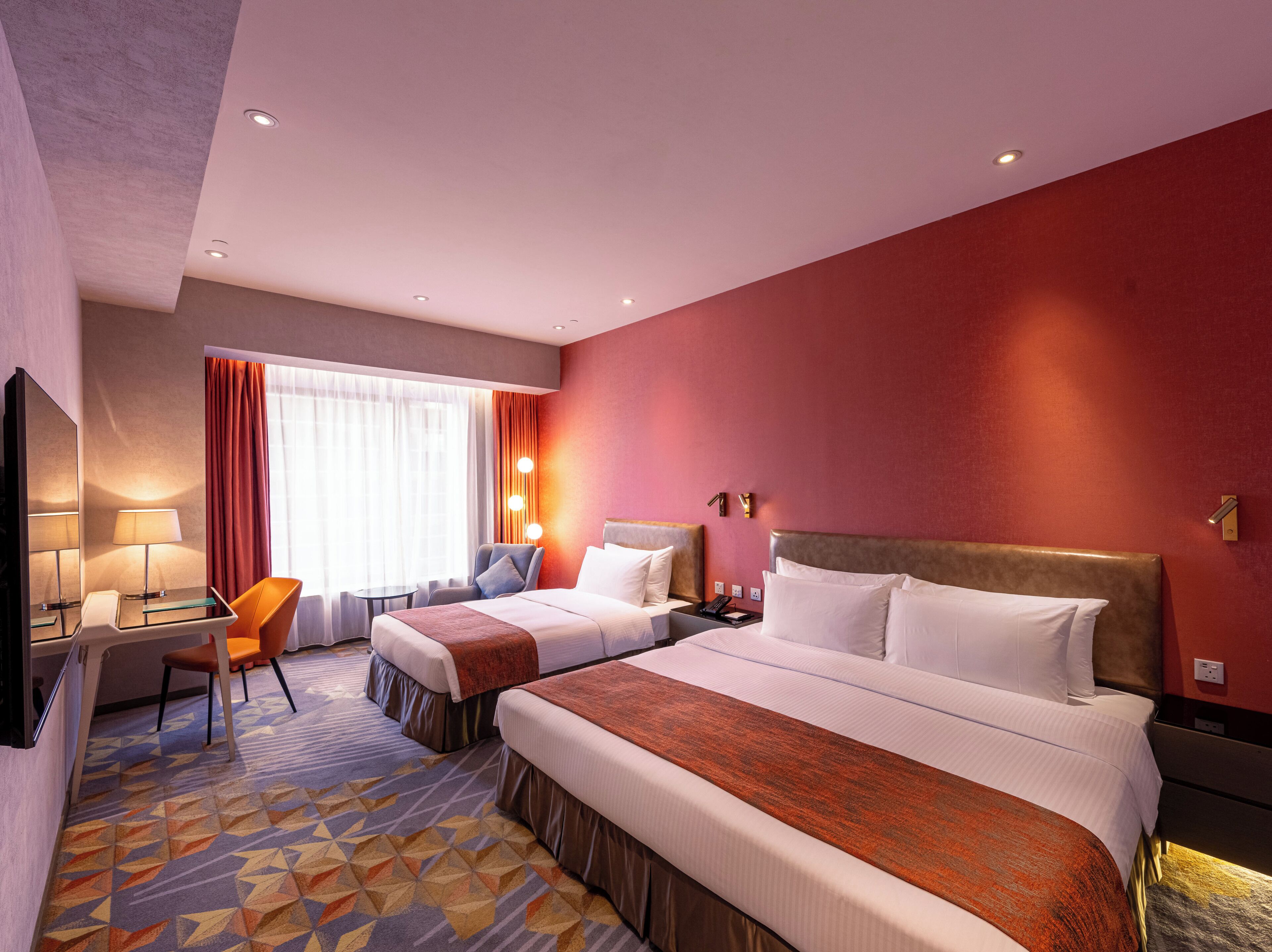A hotel room with two beds, a desk with a lamp, a chair, and a view of the outdoors through a window with curtains.