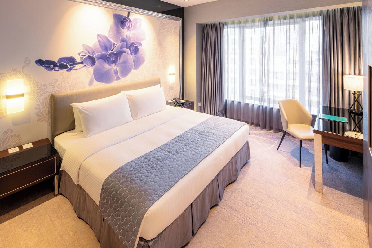A hotel room with a large bed, a desk with a chair, a lamp, and a view of the outdoors through the window.