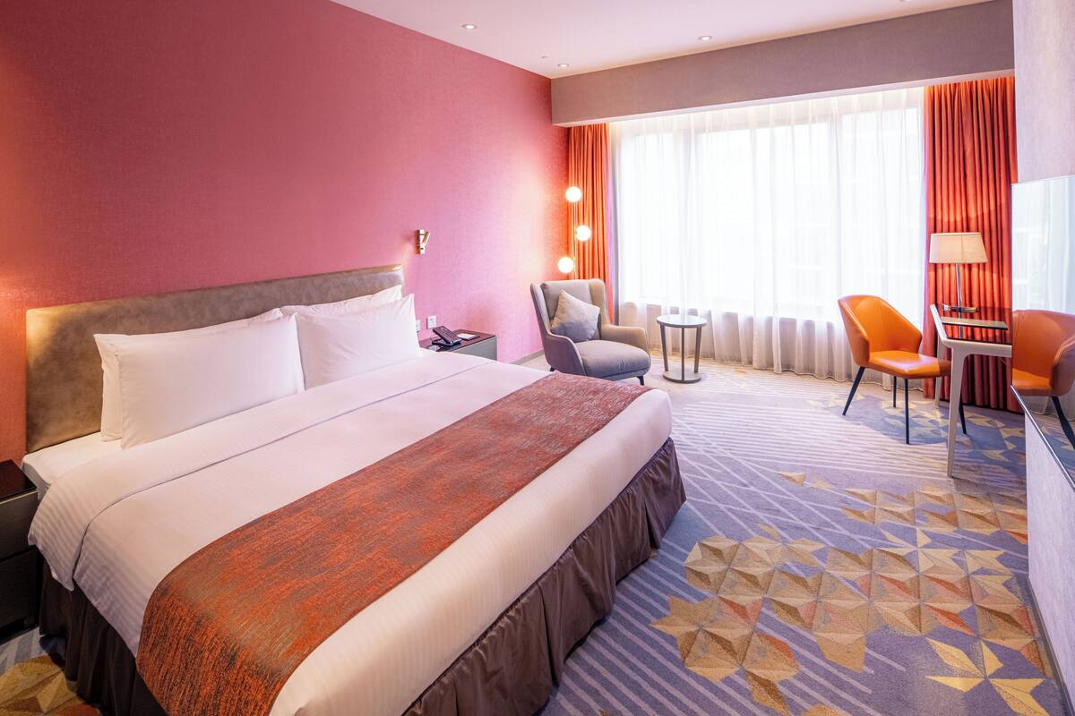 A hotel room with a large bed, a desk, two chairs, a small table, and a window with curtains.