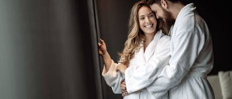 A couple in white bathrobes embracing in a modern, minimalistic room with a dark wall and a neutral-toned sofa.