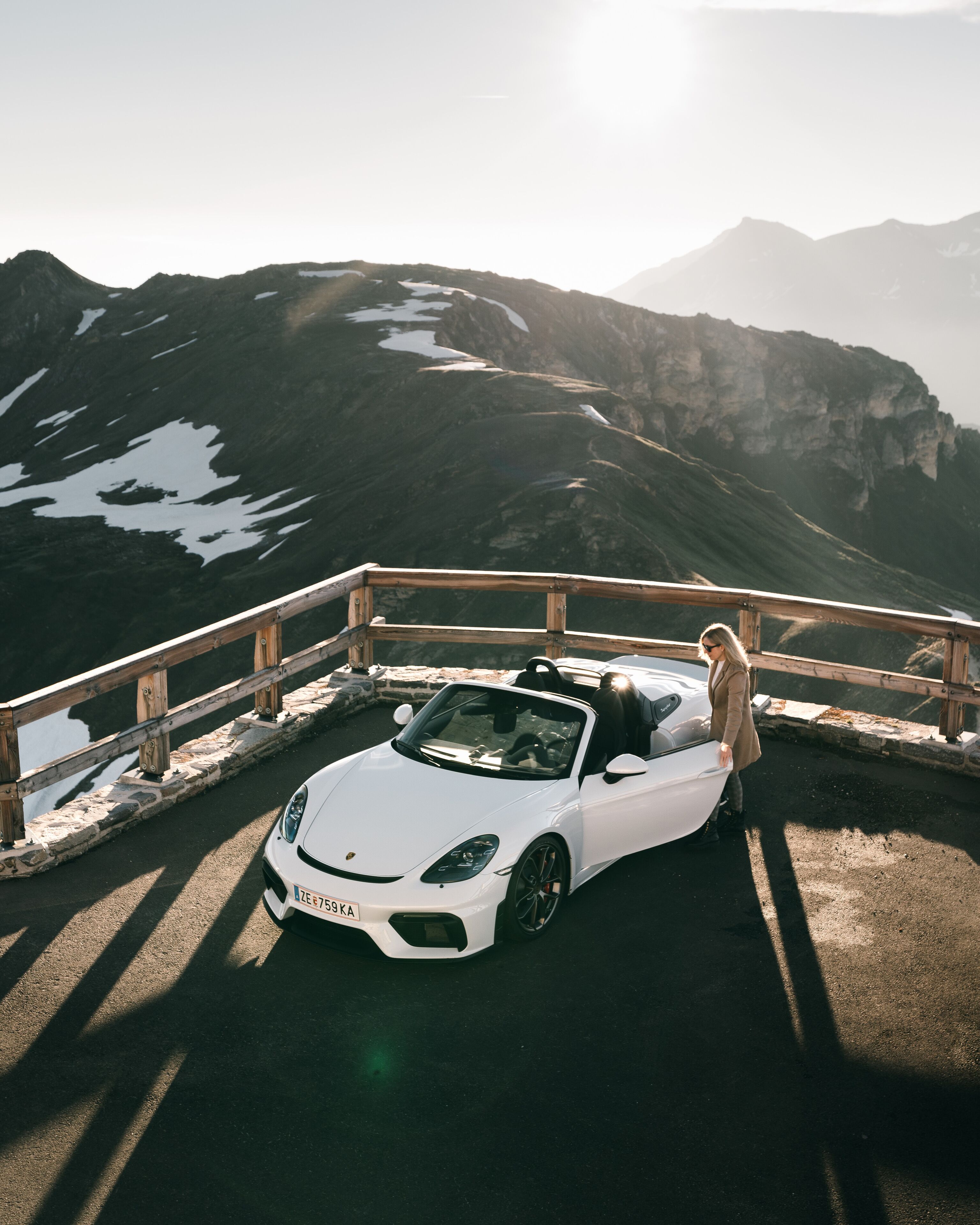 Un coche descapotable blanco estacionado en una carretera con baranda de madera, con vistas a un paisaje montañoso con parches de nieve.
